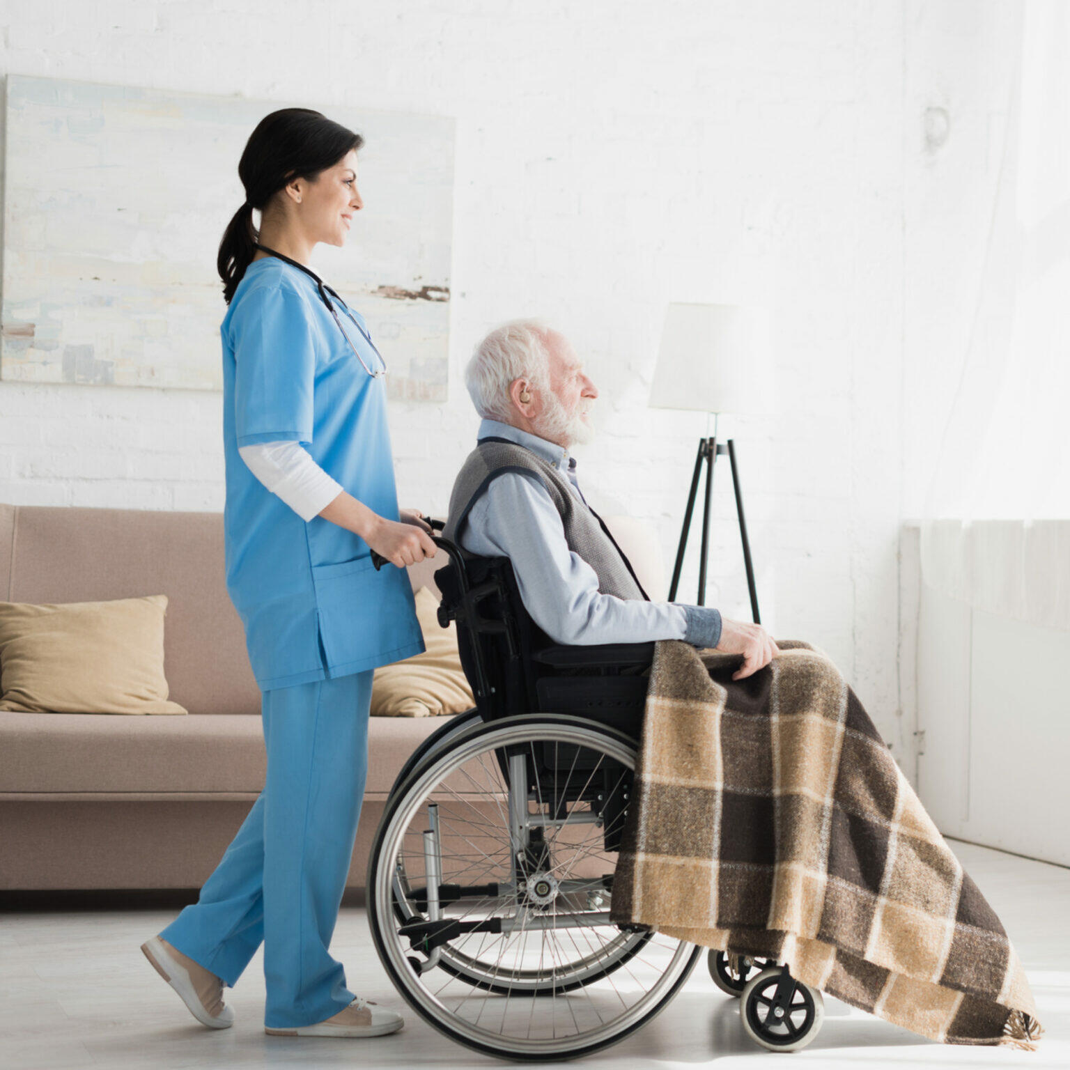 Um cuidador de uniforme azul empurra um homem idoso em uma cadeira de rodas. O homem tem um cobertor sobre as pernas e eles estão em uma sala clara e bem iluminada.