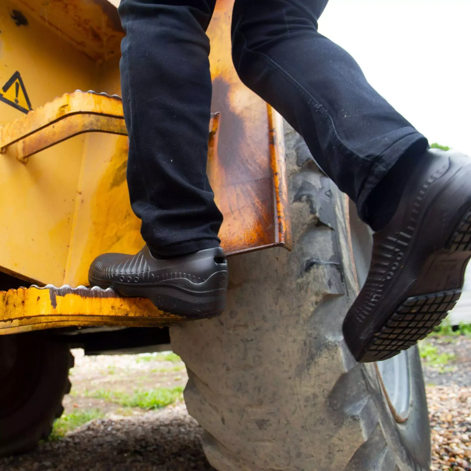 Une personne portant des chaussures noires monte dans un grand véhicule industriel jaune avec un marchepied en métal usé.