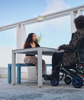 Dos mujeres sentadas a una mesa en una silla de ruedas eléctrica Q50r Carbon.