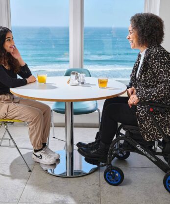 Dos mujeres sentadas a una mesa en una silla de ruedas eléctrica Q50r Carbon.