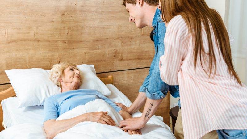 A woman lying in a hospital bed with two people standing next to her, one of them holding her hand and the other leaning over and looking at her.