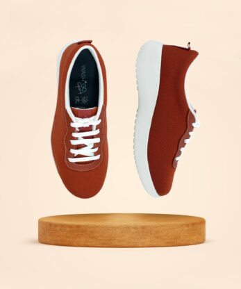 Dos zapatillas Wash'Go Sport Amber de color marrón, con cordones y suela blancos, están suspendidas sobre una plataforma de madera sobre un fondo beige.