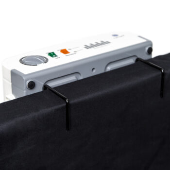 Primer plano de un Colchón Tubular Antidecúbito con Compresor acoplado en tejido negro. El dispositivo blanco, parte esencial del sistema antidecúbito, tiene un dial, un botón verde, una etiqueta naranja y un gráfico de barras. Dos ganchos negros sujetan la tela.